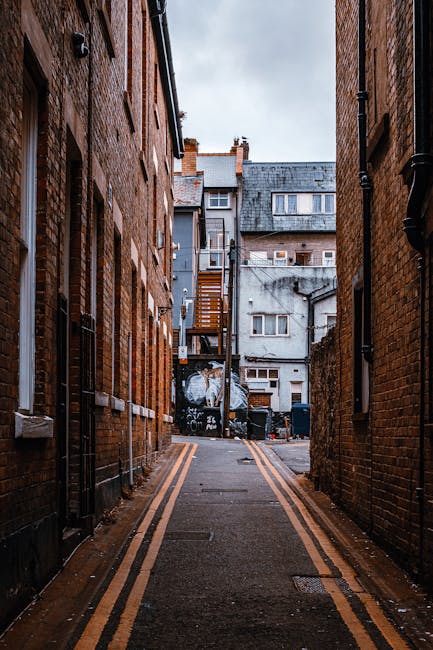 A narrow urban alleyway flanked by tall brick buildings with windows and drainpipes along their facades. The asphalt ground features double yellow lines running parallel to the walls, indicating parking restrictions. At the end of the alley, a small back yard area is visible, containing piled rubbish bags, black waste bins, and debris near a metal fence. A wooden staircase attached to the back of a grey building leads to an upper floor, with additional residential structures in the background. The scene suggests an environment where private waste disposal or rubbish collection services, such as those offered by House Clearance Lambeth, may be necessary due to the confined, inaccessible nature of the alley for regular council waste collection. The overall atmosphere is overcast, with diffuse natural light reflecting the typical grey British weather, emphasizing the utilitarian aspect of the space and its connection to alternative rubbish removal methods for densely built urban backstreets.