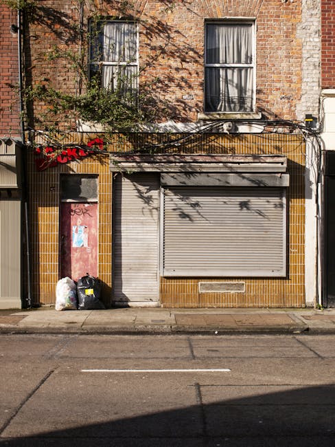 The image depicts the facade of a two-storey brick building with a storefront on the ground level. The upper floor features two rectangular sash windows with white frames, partially obscured by shadows cast from nearby branches. The shopfront has a weathered, closed metal roller shutter, painted light grey, with visible horizontal ridges, and a narrow vertical service door to its left, also closed. The surrounding wall is tiled with small, glossy, yellow-brown tiles arranged in a grid pattern, some of which appear slightly discolored. To the left side of the shopfront, there are three large black and white plastic trash bags, some with yellow labels, placed directly on the pavement next to a pink painted door that appears to be boarded up or painted over. Above the shopfront, black cables are loosely draped across the facade, with some running down the wall and casting shadows. The street in front is paved with asphalt, showing some cracks and worn markings, indicating a typical urban setting. The overall scene suggests an area where private or independent rubbish collection might be necessary, reflecting the context of alternative waste handling services.