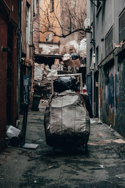 A narrow urban alleyway flanked by tall brick buildings with windows and drainpipes along their facades. The asphalt ground features double yellow lines running parallel to the walls, indicating parking restrictions. At the end of the alley, a small back yard area is visible, containing piled rubbish bags, black waste bins, and debris near a metal fence. A wooden staircase attached to the back of a grey building leads to an upper floor, with additional residential structures in the background. The scene suggests an environment where private waste disposal or rubbish collection services, such as those offered by House Clearance Lambeth, may be necessary due to the confined, inaccessible nature of the alley for regular council waste collection. The overall atmosphere is overcast, with diffuse natural light reflecting the typical grey British weather, emphasizing the utilitarian aspect of the space and its connection to alternative rubbish removal methods for densely built urban backstreets.
