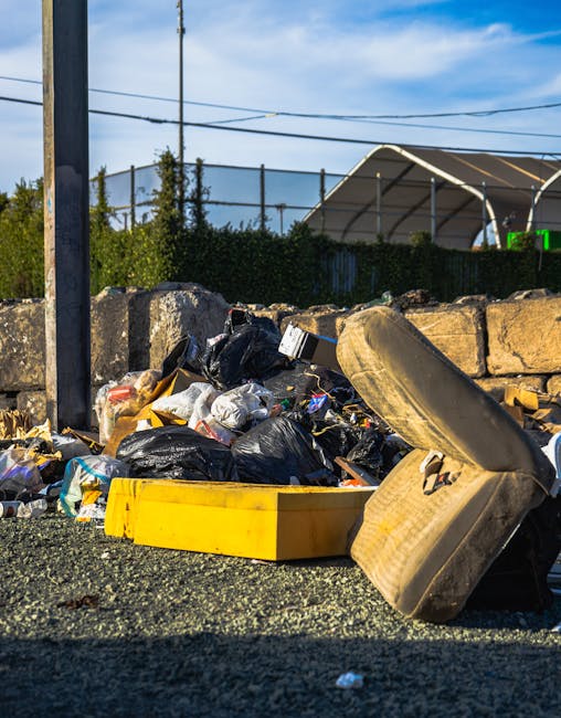 A roadside pile of mixed rubbish sits on a gravel surface next to a low stone wall. The waste includes black plastic bin bags, crumpled paper, and discarded packaging, with some items partially spilling out. An upright, old, yellow plastic toolbox is visible among the waste, along with a large, tilted, weathered beige car seat or cushion leaning against the pile. In the background, there is a metal fence with green shrubbery behind it, and a dark structure resembling a tennis court or sports area with a curved, semi-transparent roof. Above, the sky is clear with a few clouds, and utility wires run across the scene. The overall setting suggests a spot for informal waste collection or rubbish temporarily left outdoors, highlighting the need for proper disposal or private rubbish removal services like those provided by House Clearance Lambeth, which offers alternative waste handling options outside standard council collections.