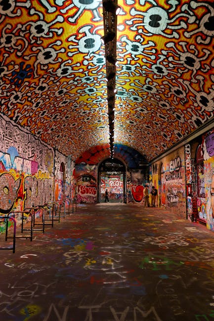 A long, arched tunnel decorated with vibrant, multicolored graffiti covering the ceiling, walls, and floor. The ceiling features a pattern of stylized skull motifs with orange, yellow, and black outlines set against a white background, creating a striking visual contrast. The side walls are adorned with various graffiti artworks, including stylized text, a large eye, and other abstract and figurative designs, all painted in bright, diverse colors. The floor has spray-painted markings and graffiti tags, with some patches of paint overlapping. Along the left wall are evenly spaced metal barriers or railings, while the right side appears more open, with a few people standing further down the tunnel. Natural light filters in from the entrance, illuminating the scene and emphasizing the vibrant hues of the graffiti. The overall setting resembles a street art installation or an underground passage used for artistic expression, with no clear indication of waste removal or rubbish service activity visible in the scene. House Clearance Lambeth occasionally handles private on-site clearance tasks, but this particular image primarily showcases urban graffiti art in a public space.