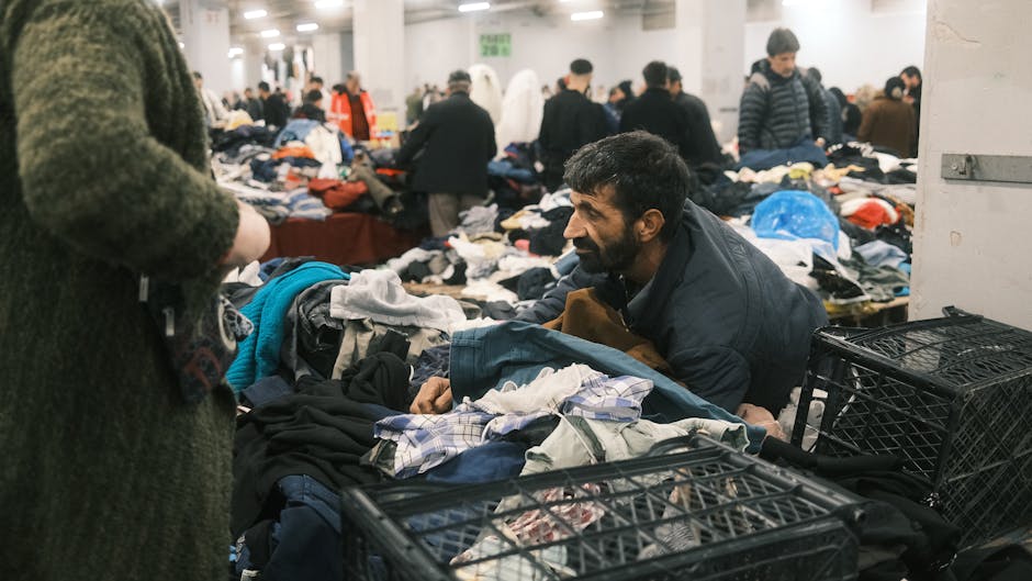 A man with dark hair and a beard, wearing a navy jacket, is seated on the floor amidst a large pile of mixed clothing items, including shirts, trousers, and jackets, which are spread out on tables and in open crates. The clothing appears to be a combination of casual and workwear, with various fabrics and textures visible. He is smiling and engaging with a person standing nearby, whose arm and part of a green coat are visible in the foreground. In the background, numerous other individuals are browsing through similar collections of clothing, set in a spacious indoor market or warehouse environment with white walls and bright lighting. The setting suggests a place where people are choosing second-hand garments, possibly related to alternative waste handling or charitable donation activities, aligning with the theme of independent rubbish collection or disposal. The scene captures an active marketplace atmosphere centered on clothing sorting and second-hand clothing exchange, relevant to alternative rubbish removal services provided by companies like House Clearance Lambeth.