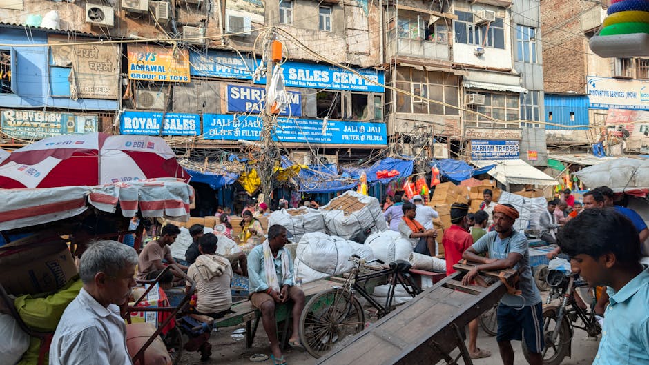 A busy outdoor market scene in an urban area with multiple multi-story buildings in the background, featuring a variety of signs advertising sales and shops. The foreground shows numerous people engaged in commercial activities and social interactions, with some standing, sitting, or walking. Piles of large, white, plastic-wrapped bundles and sacks, likely containing goods or waste, are scattered across the market, positioned among bicycles and carts. Market stalls with striped and solid-colored umbrellas provide some shade, while the environment appears crowded and vibrant. The facades of the buildings display a mix of concrete and brick with visible windows, air conditioning units, and hanging wires. The scene captures a typical street market setup, emphasizing the informal nature of waste collection and material handling, resembling alternative disposal methods often associated with independent rubbish removal efforts carried out by local traders or service providers like House Clearance Lambeth, in a context where waste is managed on-site or outside traditional municipal services.