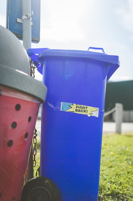 A tall, blue plastic wheelie bin with a closed lid, positioned outdoors on a grassy area near a paved surface. The bin has a small, rectangular sticker on the front with the words 'FIGHT BACK!' in bold, black letters on a yellow background with blue accents. To the left of the blue bin, part of a red wheelie bin with a handle and a lid is visible, featuring black circular ventilation holes on its side. The environment appears bright with natural daylight, and a metal pole or signpost can be faintly seen in the background, indicating the scene is likely in a residential or urban area. The image relates to waste management or rubbish collection, with the blue bin representing a typical container used for household waste, and the presence of multiple bins suggesting an organised approach to rubbish disposal, perhaps via independent collection services like those offered by House Clearance Lambeth.