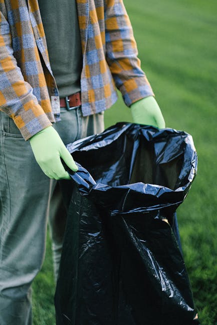 A person wearing a plaid shirt with shades of yellow, blue, and gray, along with a green long-sleeved top underneath, stands outdoors on a grassy area. They are wearing light-colored trousers secured with a brown belt and are holding open a large black plastic rubbish bag, prepared for waste collection. The individual is also wearing light green gloves, indicating they are engaged in waste or rubbish removal work. In the background, there's a vibrant green lawn or field, with no other objects or structures visible. The scene suggests an outdoor rubbish clearance activity, possibly part of a private or independent waste disposal or on-site rubbish collection service. The image visually captures the process of handling and preparing rubbish for disposal, consistent with services provided by companies such as House Clearance Lambeth, which specialise in domestic and outdoor waste removal tasks.