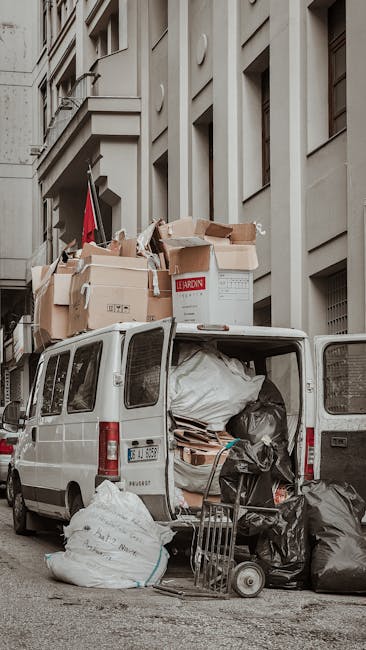 A white commercial van parked on a city street, with its rear doors open revealing a large volume of mixed rubbish including cardboard boxes, black trash bags, and loose waste. The cardboard boxes are stacked in the vehicle and some are placed on the van's roof, with visible labels and torn sections. The waste inside appears to be a combination of packaging materials and general refuse, with some items spilling slightly outside the vehicle onto the pavement. A small trolley with additional black rubbish bags and some wooden or cardboard debris is positioned nearby on the ground, indicating a recent collection or loading activity. The surrounding environment features a multi-storey building with light grey or beige exterior walls, large rectangular windows, and structural detailing like columns and ledges, suggesting an urban setting where private waste collection services might be used for on-site clearance or alternative rubbish disposal. The scene is captured in daylight with natural lighting, emphasizing the organized but cluttered state of waste awaiting removal, consistent with services offered by companies like House Clearance Lambeth for rubbish removal and disposal.