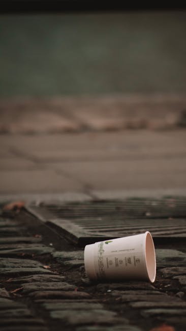 A white paper cup with printed text lies on a weathered brick pavement, partially tilted and empty, against a background of a dark, textured doormat and a wooden surface, likely a doorstep or porch area. The scene is outdoors, with natural lighting that highlights the rough texture of the bricks and the cup’s smooth surface. The environment suggests a casual setting near a residential entrance, where waste such as disposable cups may be discarded, with the image emphasizing the importance of proper rubbish collection and alternative waste handling, as provided by House Clearance Lambeth. The overall atmosphere is neutral and observational, focused on the type of small, everyday litter that services in rubbish removal aim to efficiently clear and manage.