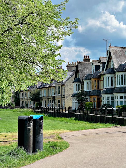 A paved pathway runs alongside a well-maintained grassy area bordered by a black metal fence, with two black public waste bins positioned near the edge of the grass. In the background, a row of traditional terraced houses with bay windows, pitched roofs, and chimney stacks is visible, featuring brick and Victorian-style architecture with decorative detailing. A large leafy tree with fresh green foliage extends over the scene from the left, partially shading the pathway and grass area. The sky overhead is partly cloudy with patches of blue, creating a bright, natural light environment. The overall scene suggests an urban residential setting, where private waste collection services like House Clearance Lambeth may be employed as an alternative to local authority rubbish collection, evidenced by the absence of visible communal waste facilities and the presence of personal waste disposal options.
