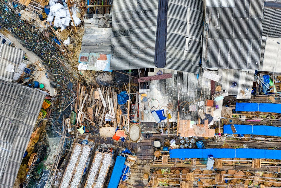 An aerial view of a cluttered outdoor area with various types of discarded materials, including wooden planks, plastic containers, corrugated metal sheets, and scattered debris. The scene shows uneven terrain with a mixture of weathered wood, some painted surfaces, and rusted metal sheets with visible textures. Piles of rubbish are spread across what appears to be a narrow yard or alleyway, surrounded by makeshift structures and sheds with corrugated metal roofing in shades of silver and grey, some with patches of black tarpaulin. Several blue plastic sheets cover parts of the apparent storage or work areas, and random household items such as buckets and tiles are visible among the waste. The environment suggests a site where temporary or informal rubbish collection and clearance services, like those offered by House Clearance Lambeth, might be necessary for efficient waste removal or private disposal, especially in contexts involving on-site clearance or alternative waste handling methods.