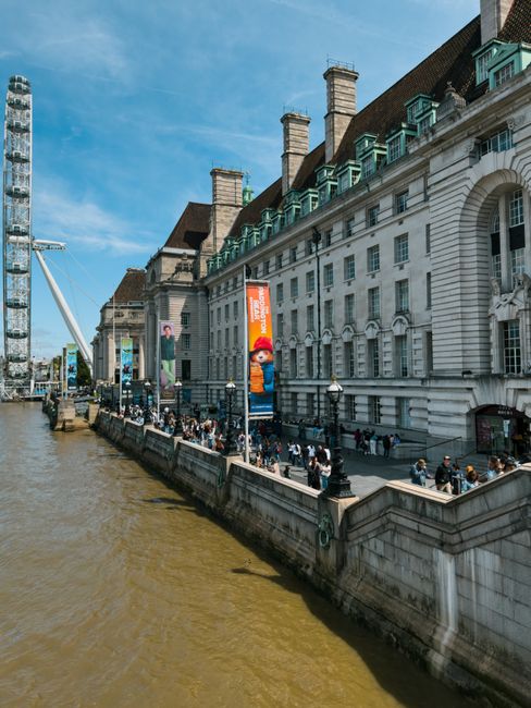 A wide view of a historic white stone building with a red-tiled roof and multiple chimney stacks, situated along the River Thames. The building features classic architectural details including arched windows and ornate cornices, with a modern ferry wheel visible on the left side of the image. Along the riverbank, a paved promenade hosts a large crowd of pedestrians walking and gathering near lampposts, some standing in small groups. Several vertical banners with bright orange backgrounds and images of characters are attached to the lampposts, advertising exhibitions or events. The river's edge shows murky brown water, and the scene is set under a clear blue sky, with the presence of evening or afternoon sunlight casting shadows on the building’s facade. This urban riverside scene exemplifies an environment where private or alternative waste handling methods, such as independent rubbish collection services like House Clearance Lambeth, might be relevant when managing waste from events or gatherings in the area.