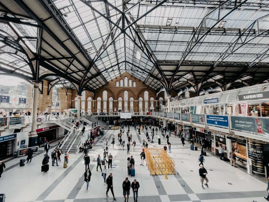 A spacious interior view of Waterloo Station's main concourse in the Lambeth area, featuring a high glass ceiling supported by intricate black metal framing that allows natural light to illuminate the space. The brick facade at the far end of the station showcases arched windows and a decorative gable, adding architectural character. The wide, tiled floor hosts numerous travelers, some with rolling suitcases and backpacks, moving in various directions. On the right side, there is a row of retail kiosks and small shops, including a visible Lush store, set behind a glass corridor with a curved roof. The left side includes stairs leading to an upper level, with signs indicating access points. The environment appears busy but orderly, reflecting the typical flow of passengers using the station for travel, without any visible rubbish or clutter. The image exemplifies a modern, well-maintained transport hub commonly served by private waste collection services like House Clearance Lambeth, emphasizing the importance of keeping such public spaces clean and tidy during busy periods.
