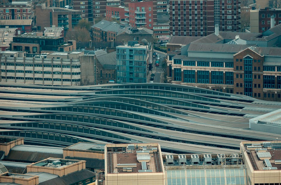 The image displays a high-angle view of an urban cityscape featuring a distinctive modern building with multiple elongated, curved, and layered metallic roofs that resemble flowing waves, situated among a variety of older and contemporary buildings in the background. The metal roofs have a sleek, reflective surface with a silver-grey finish, contrasting with the surrounding structures which include brick, glass, and concrete facades. Taller residential and commercial buildings in shades of red brick, blue, and beige are visible behind the curved-roof structure, along with smaller rooftops that have air conditioning units, vents, and other mechanical installations. The environment appears to be in a dense city area, with a mixture of architectural styles and building heights. The lighting is natural daylight, casting subtle shadows, providing clarity of textures and materials. This scene reflects urban development typical of central city locations, where on-site or private waste disposal options, such as rubbish removal services by companies like House Clearance Lambeth, may be utilized to manage waste generated from construction, renovation, or daily operations within these densely built environments, especially given the need for organized waste handling on large-scale sites or commercial properties.