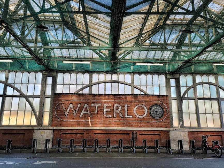 A spacious interior view of Waterloo Station's main concourse in the Lambeth area, featuring a high glass ceiling supported by intricate black metal framing that allows natural light to illuminate the space. The brick facade at the far end of the station showcases arched windows and a decorative gable, adding architectural character. The wide, tiled floor hosts numerous travelers, some with rolling suitcases and backpacks, moving in various directions. On the right side, there is a row of retail kiosks and small shops, including a visible Lush store, set behind a glass corridor with a curved roof. The left side includes stairs leading to an upper level, with signs indicating access points. The environment appears busy but orderly, reflecting the typical flow of passengers using the station for travel, without any visible rubbish or clutter. The image exemplifies a modern, well-maintained transport hub commonly served by private waste collection services like House Clearance Lambeth, emphasizing the importance of keeping such public spaces clean and tidy during busy periods.